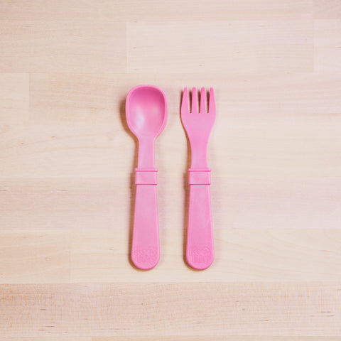 A pink plastic Re-Play Utensil Set from REPLAY, featuring a spoon and fork laid side by side on a light wooden surface, makes the perfect toddler utensils. They're made from recycled plastic and are dishwasher safe.