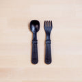 On a light wooden surface, a combination spoon/spatula and fork/spatula in sleek black from the REPLAY brand are displayed. Both utensils, crafted from recycled plastic, features wide handles and flat bases with "Re:Play" imprinted on them. This Re-Play Utensil Set is dishwasher-safe and ideal for your toddler's mealtime fun.