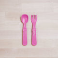 A pair of Re-Play Utensils from REPLAY are placed side by side on a light wood surface. The left utensil is a spoon, and the right utensil is a fork. Both utensils, designed for toddlers, are made of recycled plastic, featuring wide handles and being dishwasher safe.