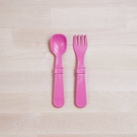 A pair of Re-Play Utensils from REPLAY are placed side by side on a light wood surface. The left utensil is a spoon, and the right utensil is a fork. Both utensils, designed for toddlers, are made of recycled plastic, featuring wide handles and being dishwasher safe.