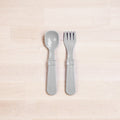 The Re-Play Utensil Set by REPLAY, featuring a gray plastic spoon and fork made from recycled materials, lie parallel to each other on a light-colored wooden surface. The toddler utensils boast a simple design with wide handles and a slightly glossy finish.