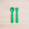 A Re-Play Utensil Set, featuring a green plastic spoon and fork designed for young children, lies side by side on a light wooden surface. Made from recycled plastic, the handles are wide and smooth, with the brand name "REPLAY" embossed near the bottom. These toddler utensils are dishwasher safe for easy cleaning.