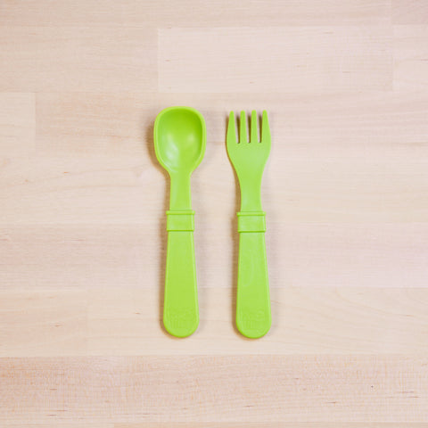 A Re-Play Utensil Set by REPLAY, featuring a green plastic children's spoon and fork made from recycled materials, are placed side by side on a light wooden surface. Both utensils boast wide handles designed for easy gripping by small hands and are dishwasher safe.