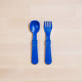 Two blue toddler utensils from the REPLAY Re-Play Utensil Set are placed on a light wooden surface. The utensil on the left is a spoon, and the one on the right is a spork fork. Both made from recycled plastic, with broad handles and are positioned parallel to each other. Plus, they're dishwasher safe for easy cleaning.