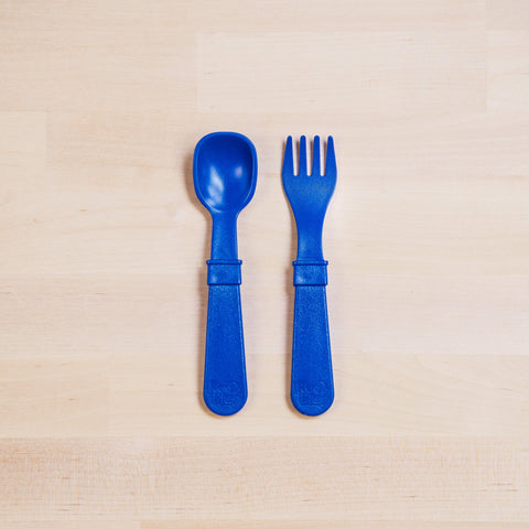 Two blue toddler utensils from the REPLAY Re-Play Utensil Set are placed on a light wooden surface. The utensil on the left is a spoon, and the one on the right is a spork fork. Both made from recycled plastic, with broad handles and are positioned parallel to each other. Plus, they're dishwasher safe for easy cleaning.