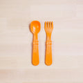 The Re-Play Utensil Set from REPLAY, crafted from recycled plastic, includes a bright orange spoon and fork. These utensils are neatly placed on a light wooden surface showcasing a natural grain pattern. Their thick handles suggest durability and ease of grip, indicating they might be designed for toddlers.