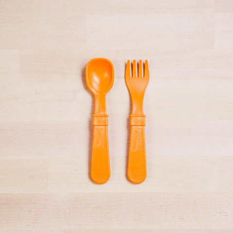 The Re-Play Utensil Set from REPLAY, crafted from recycled plastic, includes a bright orange spoon and fork. These utensils are neatly placed on a light wooden surface showcasing a natural grain pattern. Their thick handles suggest durability and ease of grip, indicating they might be designed for toddlers.