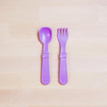 A purple Re-Play Utensil Set from REPLAY, made from recycled plastic, lies side by side on a light wooden surface. These dishwasher-safe toddler spoon and fork have wide handles and rounded edges, designed for small children to use safely.