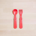 The Re-Play Utensil Set by REPLAY, featuring a red plastic toddler spoon and fork made from recycled materials, is positioned side by side on a light wooden surface. The utensils boast chunky handles for easy gripping, and the fork is designed with wide, rounded prongs. Additionally, they are dishwasher safe for added convenience.