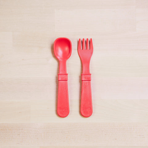 The Re-Play Utensil Set by REPLAY, featuring a red plastic toddler spoon and fork made from recycled materials, is positioned side by side on a light wooden surface. The utensils boast chunky handles for easy gripping, and the fork is designed with wide, rounded prongs. Additionally, they are dishwasher safe for added convenience.