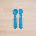 A Re-Play Utensil Set by REPLAY, consisting of a blue plastic toddler spoon and fork made from recycled materials, is displayed side by side on a light wood surface with the spoon on the left and the fork on the right. Both dishwasher-safe utensils feature thick handles for easy gripping.