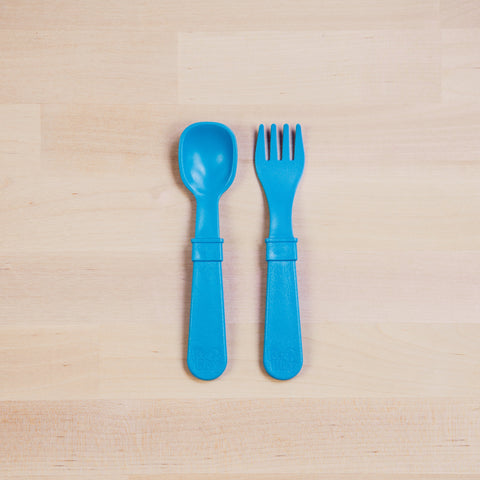 A Re-Play Utensil Set by REPLAY, consisting of a blue plastic toddler spoon and fork made from recycled materials, is displayed side by side on a light wood surface with the spoon on the left and the fork on the right. Both dishwasher-safe utensils feature thick handles for easy gripping.