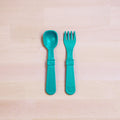 A pair of Re-Play Utensil Set, crafted from recycled green plastic and featuring a spoon and fork, is placed side by side on a light wooden surface. These REPLAY toddler utensils boast thick handles and a simple, solid design that are perfect for little hands and dishwasher safe for easy cleaning.