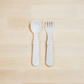 A Re-Play Utensil Set by REPLAY, consisting of a pair of white, dishwasher-safe toddler utensils—a spoon and fork—is neatly placed on a light wooden surface. Made from recycled plastic, they feature matching handles with a slightly textured grip. The background consists of light-colored wooden planks.