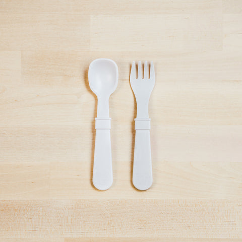 A Re-Play Utensil Set by REPLAY, consisting of a pair of white, dishwasher-safe toddler utensils—a spoon and fork—is neatly placed on a light wooden surface. Made from recycled plastic, they feature matching handles with a slightly textured grip. The background consists of light-colored wooden planks.