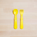 A Re-Play Utensil Set by REPLAY, consisting of a yellow plastic toddler spoon and fork, placed side by side on a light wooden surface. Made from recycled plastic and dishwasher safe.