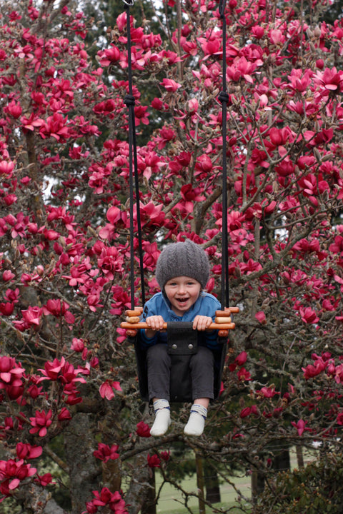 A young child wearing a gray knit hat and blue jacket joyfully swings from a SOLVEJ Baby Toddler Swing - Slate Grey by SolveJ, set against a backdrop of vibrant pink flowering trees. The child's expression is one of delight, with a bright smile on their face.