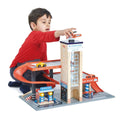 A child in a red shirt is fully immersed in imaginative play with the Tender Leaf Blue Bird Service Station from Tender Leaf. The playset features a tall blue and beige parking tower labeled "Bluebird," complete with a curved red ramp, gas station, and car wash area. The child is reaching the top level of the tower with his hand.