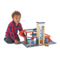 A young child clad in a red and blue checkered shirt is captivated by the Tender Leaf Blue Bird Service Station, a wooden toy parking garage set from Tender Leaf that features a ramp, elevator, gas station, car wash, and an assortment of wooden cars.