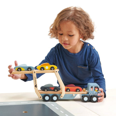 A young child with curly hair and a blue long-sleeve shirt is playing with a wooden Tender Leaf Car Transporter by Tender Leaf. The truck carries four colorful super cars: two on the lower level and two on the upper level. The child is interacting with one of the toy cars.