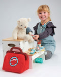 A young child with blonde hair, dressed in a blue apron dress over a dark long-sleeve shirt, is engaging in imaginative play by pretending to be a doctor. They are using the Tender Leaf Doctors Bag & Accessories and various healthcare toys to give a plush teddy bear a check-up.