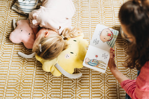 A young girl lies on a yellow plush toy with her eyes closed while a person next to her reads "The Kiss Co I got you some Kisses- board book," a charming story from The Kiss Co. The girl, adorned with a hair clip in her blonde hair, rests peacefully on the yellow patterned rug, part of the reader's red shirt and arm visible beside her.