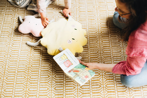 A person in a pink shirt reads "The Kiss Co I got you some Kisses" children's board book while sitting on a patterned rug. Two young children play nearby with a plush yellow cloud toy and a pink plush toy. The image depicts a cozy and playful setting for the little ones.