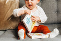 A baby with a happy expression sits on a sofa, holding The Kiss Co I got you some Kisses- board book. Another book titled "The Gift of a Cuddle" rests beside them. The little one wears an off-white top, orange pants, and white socks.