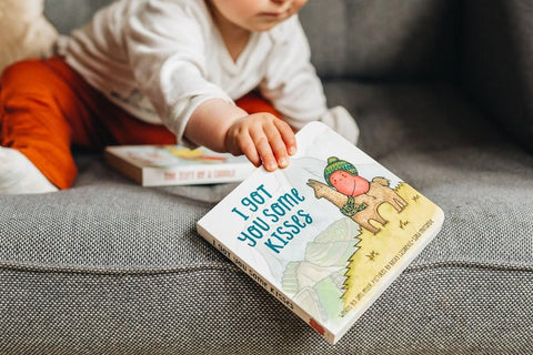 A young child in a white shirt and orange pants sits on a gray couch, reaching out to pick up "The Kiss Co I Got You Some Kisses" board book by The Kiss Co. Another classic story, ideal for little ones, is visible on the couch in the background.