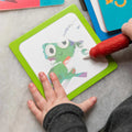 A toddler is coloring a green, cartoonish frog on a small square card from the Tiger Tribe Magic Painting ABC - Alphabet Adventures set using a red marker. The frog is partially colored, and the child's left hand is holding the card while the right hand uses the marker. Other colorful reusable Magic Painting cards from TIGER TRIBE are visible in the background.