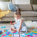 A young girl with pigtails sits on a rug in a cozy living room, surrounded by Tiger Tribe Magic Painting ABC - Alphabet Adventures cards. She holds a red marker in her left hand and points to one of the cards with her right hand. Behind her, there's a grey couch adorned with a blue cushion and a yellow throw.