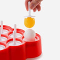 A hand holding a yellow ball on a white stick hovers over a red plastic base with multiple white pegs and depressions, one of which contains a mini pop also on a stick. This setup appears to be part of the Zoku Mini Pops by Zoku, which are BPA and phthalate-free.