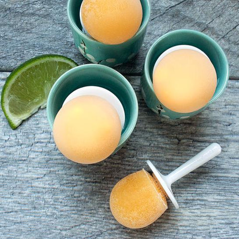 Three orange-colored frozen popsicles in decorative teal cups, made using Zoku Mini Pops molds (from the trusted brand Zoku), with one popsicle outside the cup on a plastic stick. A slice of lime is placed next to the cups on a rustic wooden surface.