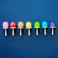 Seven mini pops are lined up against a blue background. Each pop, made with Zoku Mini Pops by the brand Zoku, features a white stick and brightly colored spherical candy arranged in a gradient from red, orange, yellow, green, light blue, blue to purple. These treats are BPA and phthalate-free.