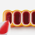 A close-up of the Zoku Mod Pops, a BPA-free, yellow four-slot popsicle mould from Zoku, being filled with red liquid from a white pitcher. The mould features four cylindrical containers arranged in a row; three are already filled to the top while the fourth is being poured into. A built-in drip guard ensures no mess during the process.